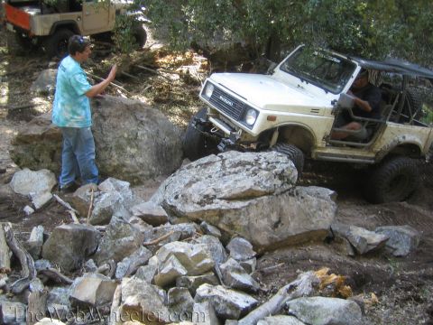 1987 Suzuki Samurai - A white 1987 Suzuki Samurai is shown off-roading over large rocks, with a person standing nearby to guide it.