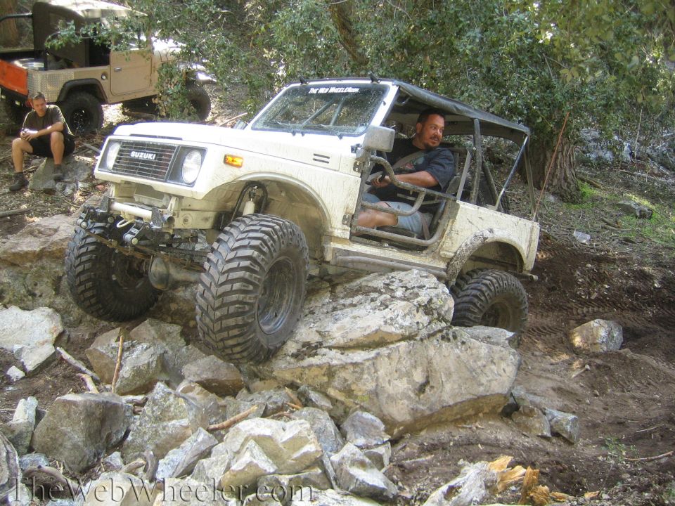 White 1987 Suzuki Samurai climbing over rocks on a trail. Another off-road vehicle is parked behind it
