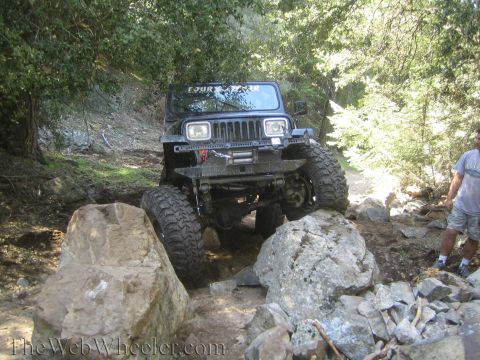 1987 Suzuki Samurai - A black Jeep Wrangler tackles a challenging off-road course, with large rocks obstructing its path.