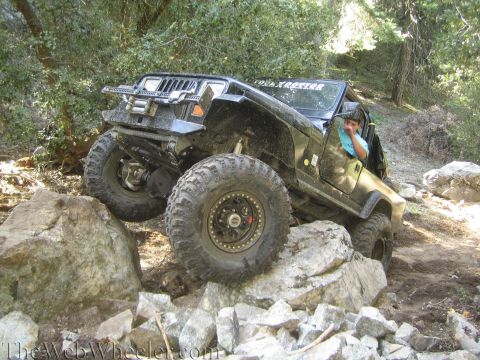 1987 Suzuki Samurai - A driver navigates a black Jeep Wrangler over a rocky obstacle on a wooded trail.