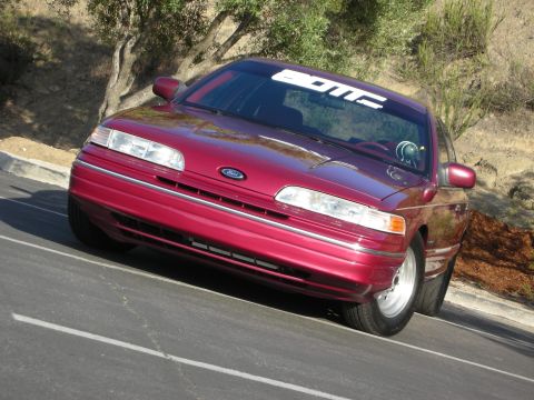 1992 Ford Crown Victoria - A red 1992 Ford Crown Victoria is parked on an asphalt surface with a tree and hillside in the background.