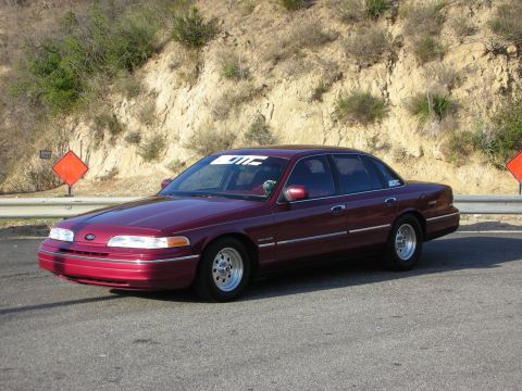 1992 Ford Crown Victoria - A maroon 1992 Ford Crown Victoria is parked on the side of a road with a hillside in the background.