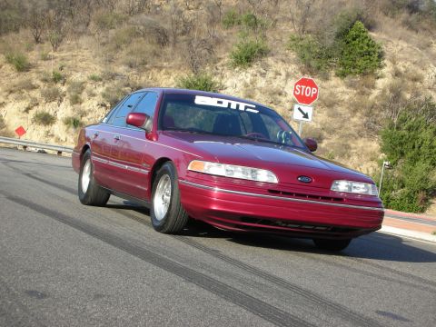1992 Ford Crown Victoria - A red 1992 Ford Crown Victoria is shown on a road with a stop sign in the background.