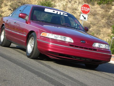 1992 Ford Crown Victoria - A red 1992 Ford Crown Victoria is shown from a low front three-quarter angle on a road with visible tire marks.