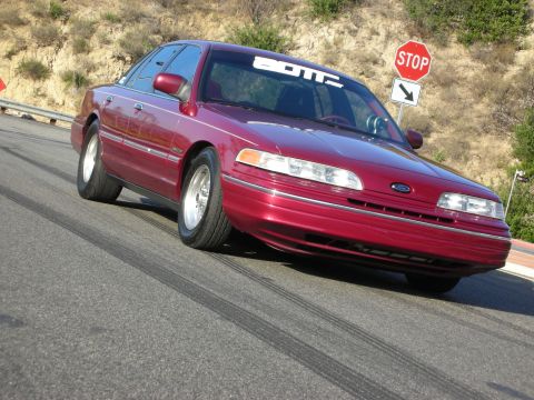 1992 Ford Crown Victoria - A red 1992 Ford Crown Victoria is shown with a banner on its windshield and tire marks on the road.