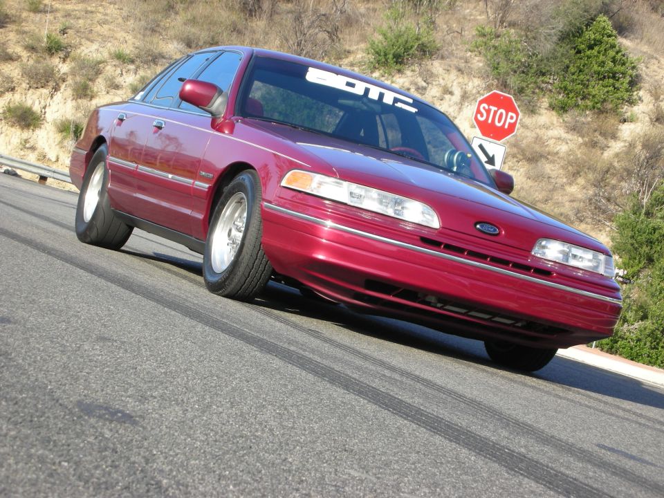 Red 1992 Ford Crown Victoria with "BOTZ" banner on windshield, driving on a road with tire marks