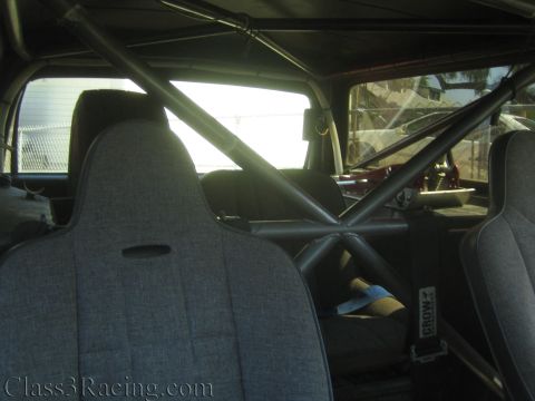 1990 Ford Bronco - interior of a stripped race truck shows gray bucket seats, a tubular roll cage and harness mounts.