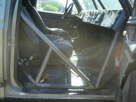 1990 Ford Bronco - Interior of a stripped truck cab with a tubular roll cage, single racing seat, dash, and a red fire extinguisher.
