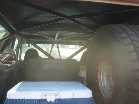 1990 Ford Bronco - Inside a vehicle's rear cargo area showing a roll cage, bench, cooler, and a large spare tire on a red beadlock wheel.