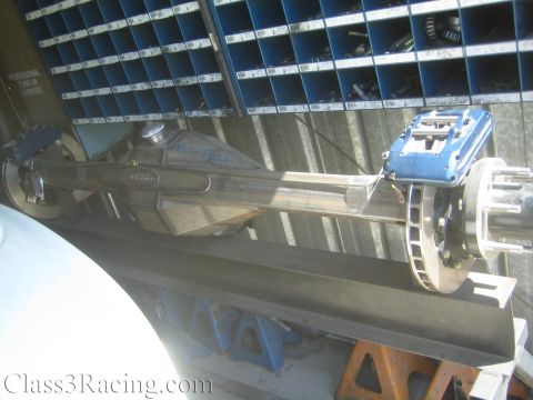 1990 Ford Bronco - fabricated rear axle assembly with a polished housing, blue brake caliper, and rotor displayed in front of a parts bin wall.