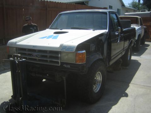 1990 Ford Bronco - black and white Ford 4x4 pickup with mismatched panels and a tube front sits on jack stands in a residential driveway.