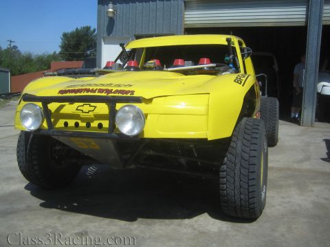 1990 Ford Bronco - yellow Chevrolet off-road race truck with fiberglass body, light bar, and bypass shocks parked by an open shop bay.