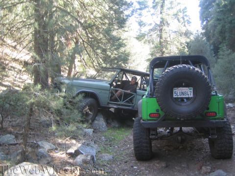 1987 Suzuki Samurai - Two off-road vehicles traverse a wooded, rocky trail, one in the foreground and another partially obscured by trees.