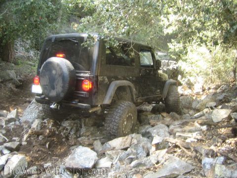 1987 Suzuki Samurai - A black Jeep Wrangler drives over a challenging rocky terrain, surrounded by trees.