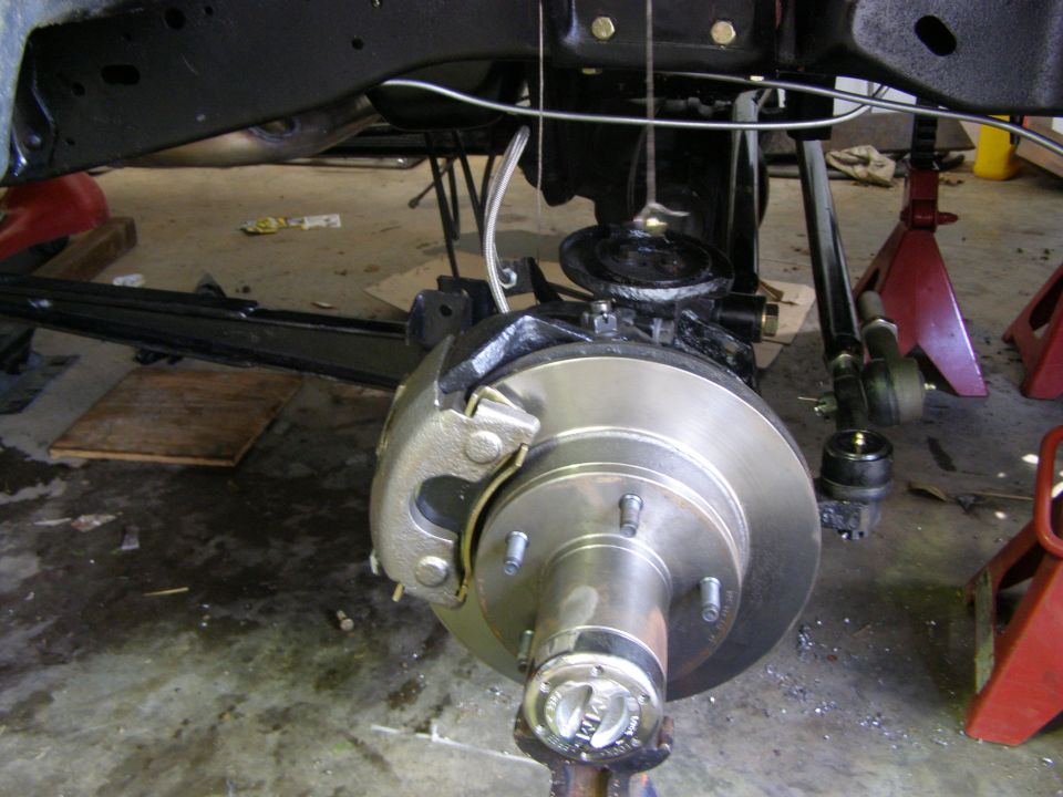 Close-up of a vehicle's front brake rotor, caliper, and suspension linkage, with a "SUPERMOTORS.NET" watermark