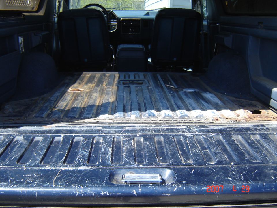 Looking over the tailgate into a Bronco's empty ribbed cargo floor with rust, toward blue front seats and dash.