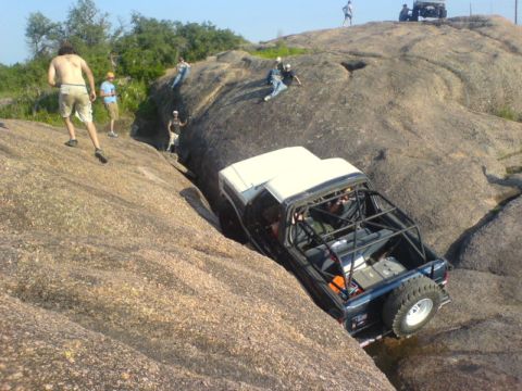 1986 Ford Bronco - An off-road vehicle attempts to climb a steep, rocky terrain with several people watching.