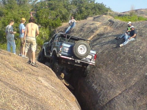1986 Ford Bronco - A modified 1986 Ford Bronco is carefully driven over large rocks, with onlookers observing the off-road challenge.