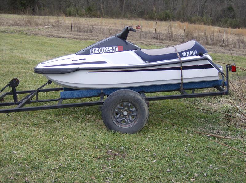 White and blue Yamaha personal watercraft strapped on a single-axle trailer in a grassy field