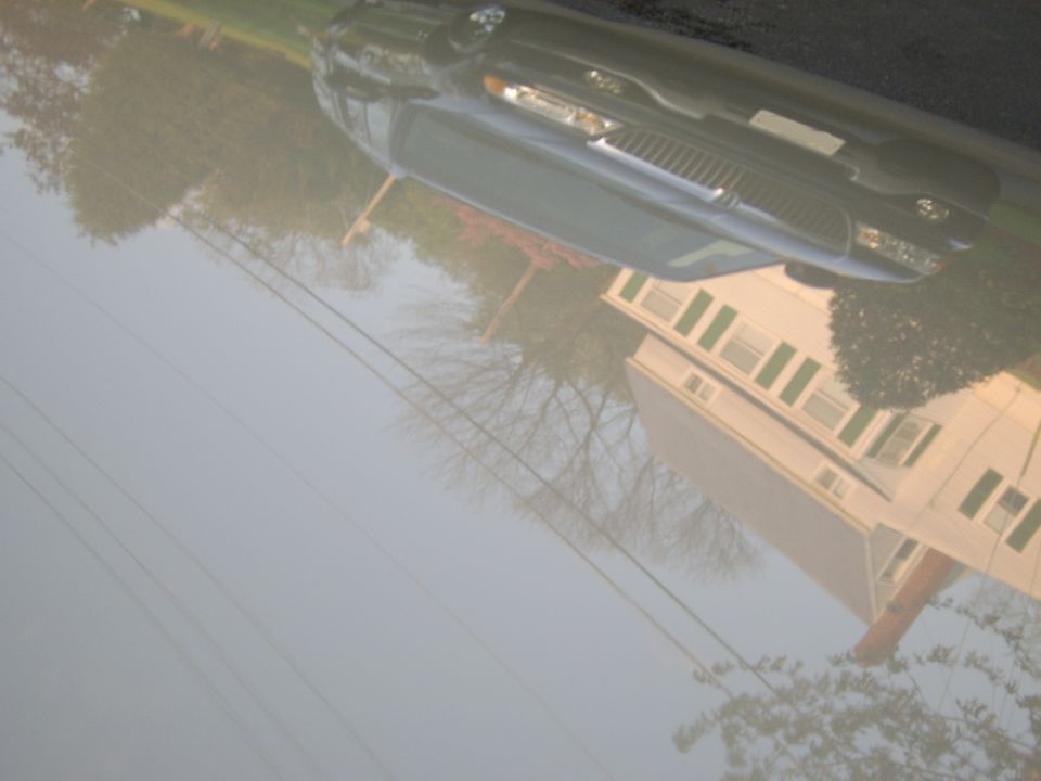 Rotated photo of dark full-size sedan front in a driveway near a house