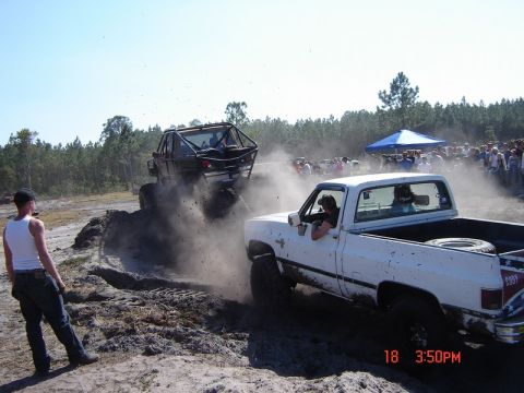 1989 Ford Bronco - Two off-road vehicles churn up dirt and dust during a competition with spectators gathered around.