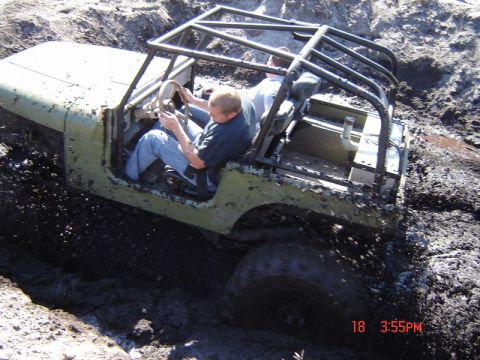 1989 Ford Bronco - An olive green off-road vehicle with a roll cage is stuck deep in mud, with two people inside.