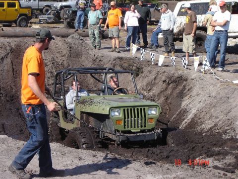 1989 Ford Bronco - A Jeep navigates a challenging mud pit during an off-road event, with spectators watching from the embankment.