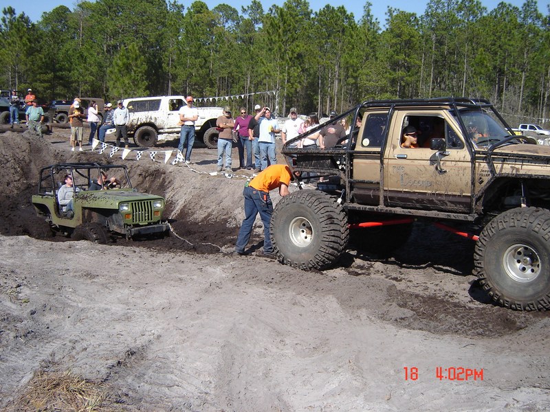 Off-road trucks and jeeps in mud pit with spectators watching. One jeep is stuck in the mud