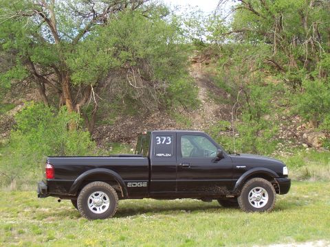 2003 Ford Ranger - A black Ford Ranger pickup truck is parked on a grassy field with trees and a dirt hill in the background.
