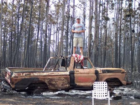 1974 Ford F-100 - A man stands atop a heavily damaged pickup truck in a forest that appears to have been affected by a fire.