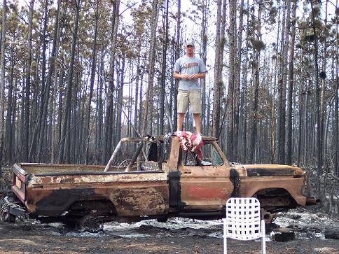 guy standing on burnt pickup truck, trees in background