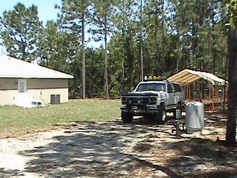 1986 Ford Bronco - A white SUV is parked in a yard next to a house, with a wooded area in the background.
