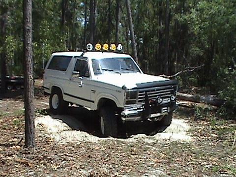 1986 Ford Bronco - A white Ford Bronco is parked in a wooded area, with a trailer visible in the background.