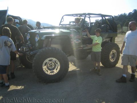 1987 Suzuki Samurai - Several people gather around a heavily modified Jeep Wrangler with oversized tires, parked outdoors.