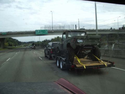 1968 Ford Bronco - A lifted olive green pickup truck is transported on a trailer on the highway.