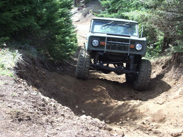 1968 Ford Bronco driving on a steep dirt trail with trees on either side