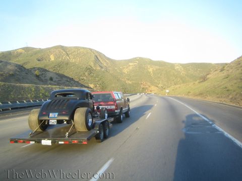 1987 Suzuki Samurai - A black hot rod is transported on a trailer behind a red pickup truck on a sunny day.