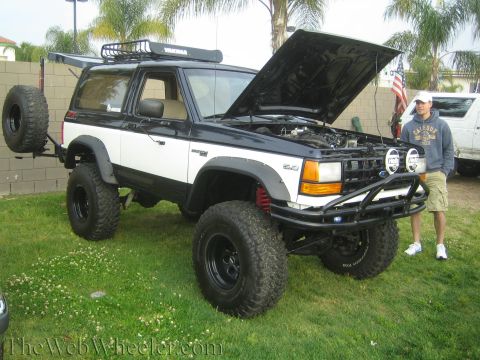 1990 Ford Bronco - lifted black and white Ford Bronco II with roof rack and open hood on grass, a man standing beside it at a show.