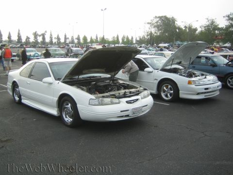 1990 Ford Bronco - Two white Ford Thunderbird coupes with hoods raised parked beside each other in a crowded car-show lot.