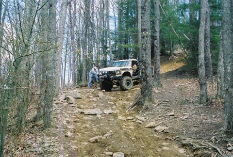 1978 Ford Bronco - A white 1978 Ford Bronco navigates a challenging, rocky off-road trail through a wooded area.