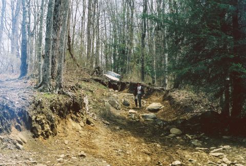 1978 Ford Bronco - A person stands on a rough dirt path as a vehicle attempts to climb a steep incline in a forest.
