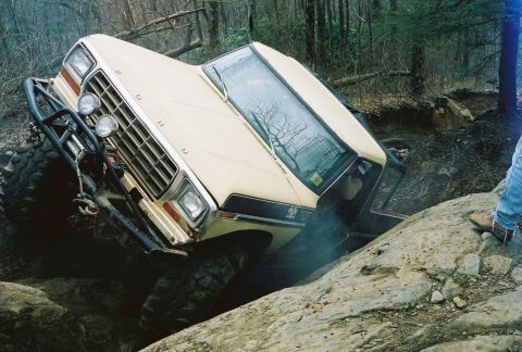 1978 Ford Bronco - A tan Ford Bronco is tilted at a steep angle, with its front tire stuck in a rocky crevice on an off-road trail. Smoke is visible near the…