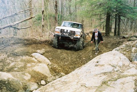 1978 Ford Bronco - A white 1978 Ford Bronco navigates a challenging rocky off-road trail with a person walking alongside.