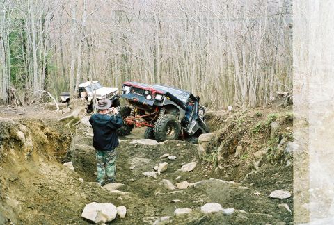 1978 Ford Bronco - A heavily modified black off-road vehicle navigates a steep, rocky ditch in a wooded area, while another white off-road vehicle is visible…