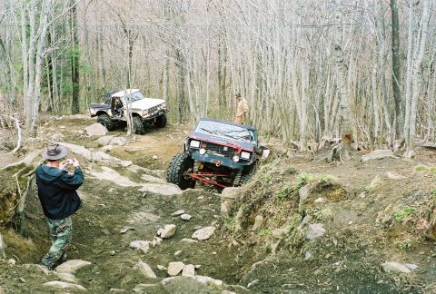 1978 Ford Bronco - Two off-road vehicles traverse a challenging, rocky path through a dense forest.