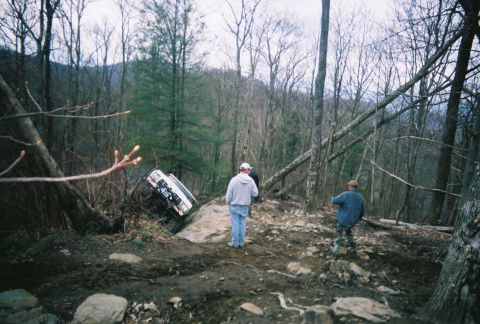 1978 Ford Bronco - An off-road vehicle is stuck on a large rock in a wooded area with two observers nearby.