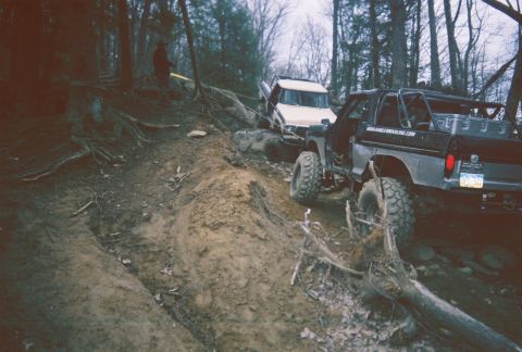 1978 Ford Bronco - Two modified trucks tackle a challenging, muddy off-road course in a wooded area.