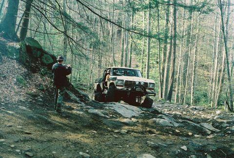 1978 Ford Bronco - A white SUV navigates a dirt road surrounded by trees.
