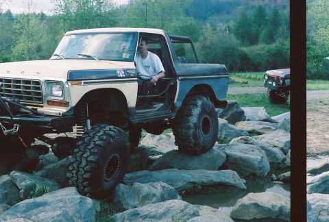 1978 Ford Bronco - A man sits in a modified 1978 Ford Bronco with oversized tires, driving over a rocky terrain.