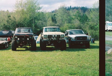 1978 Ford Bronco - Three heavily modified trucks are parked on a grassy field with trees in the background.